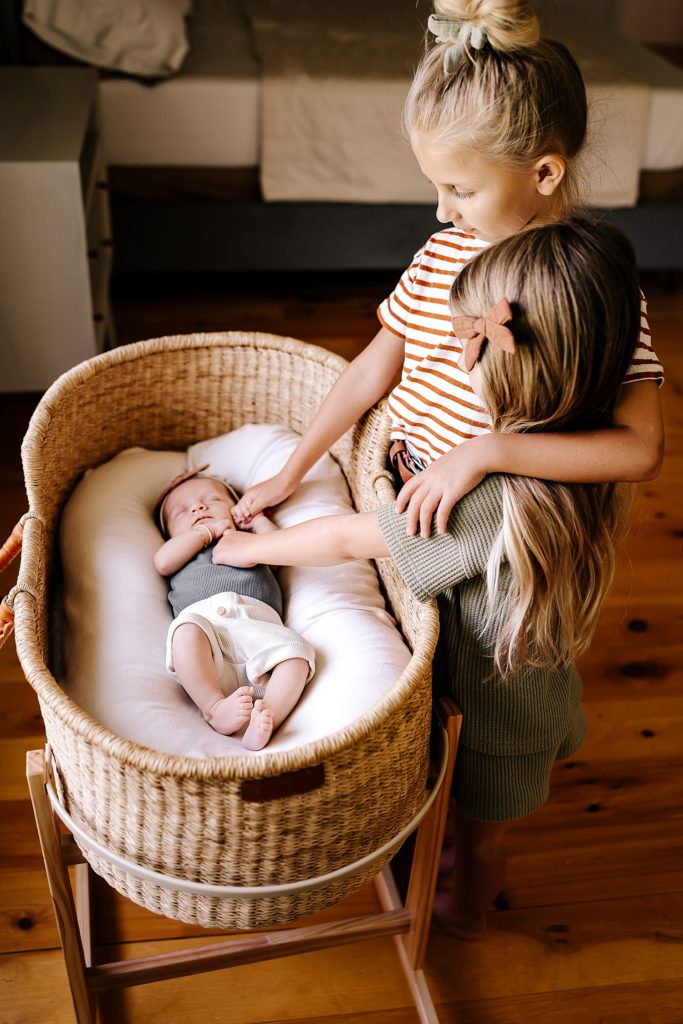 Two older sisters touching their baby sister in her bassinet. 