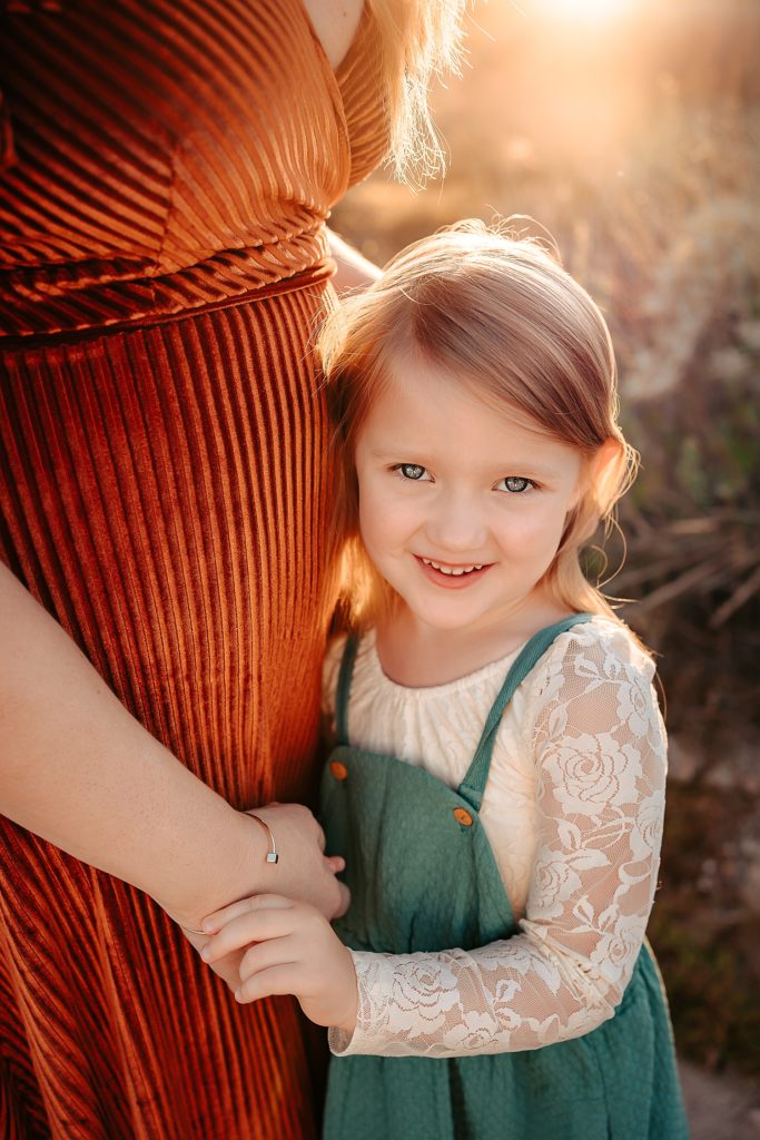Young blonde girl in green dress looking up into the camera and smiling while holding her mother's hands. 