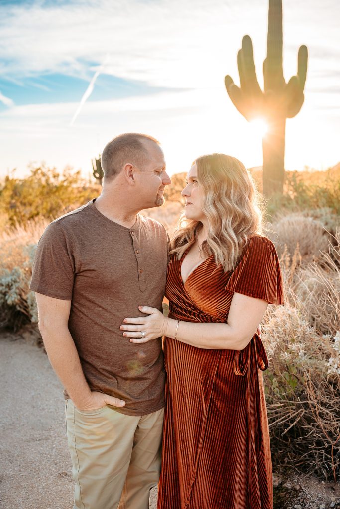 Husband and wife looking at each other and smiling with a large cactus behind them for their boho family session in Arizona.