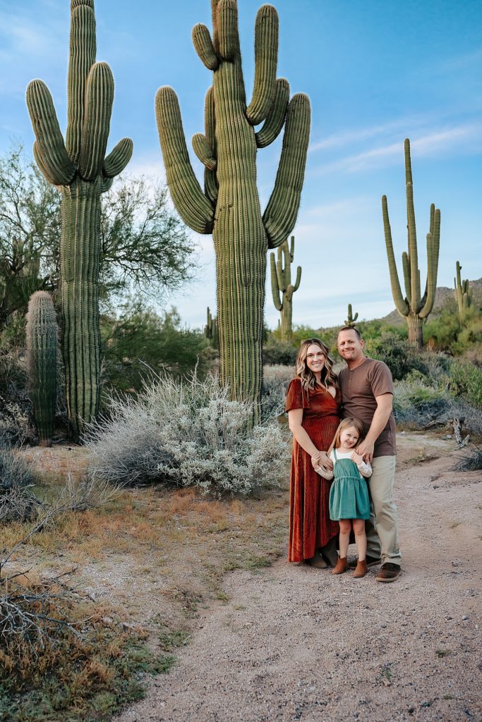 Family of three smiling at the camera surrounded by large cactuses at their boho family session in Arizona. 
