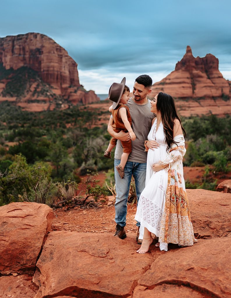 Family of three with mom in white dress, holding her pregnant belly, with the Arizona scenery behind them in vibrant colors.