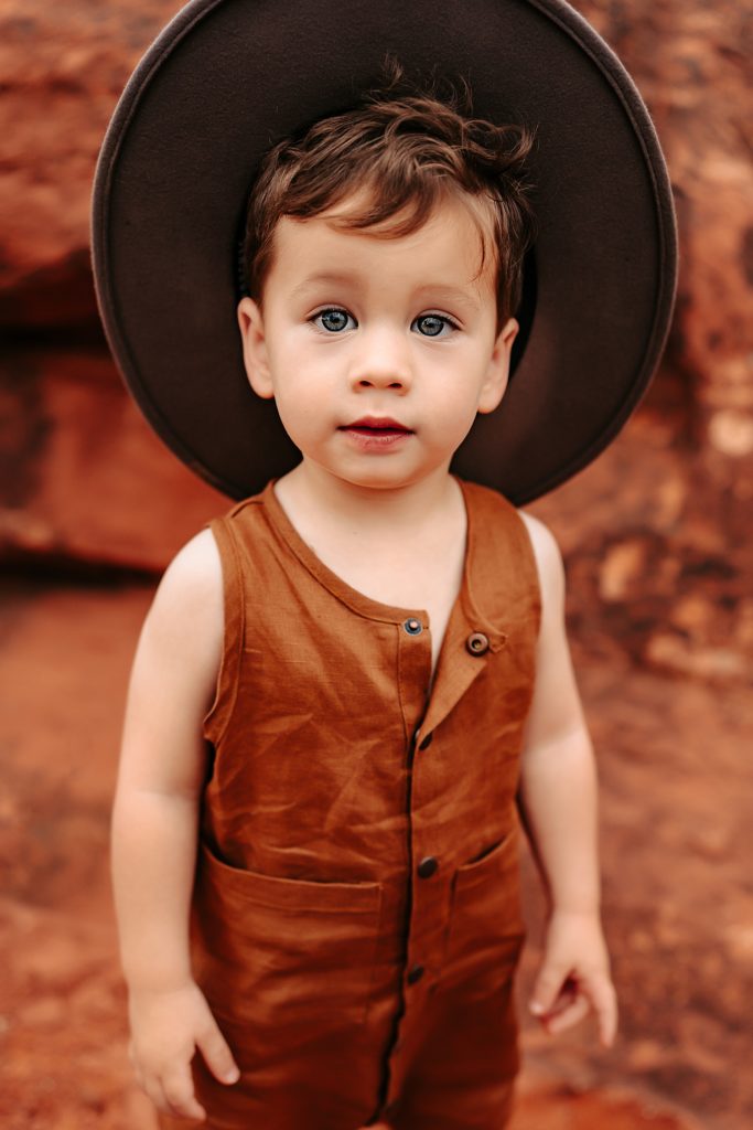 Toddler boy in brown linen jumpsuit and oversized hat looking into the camera with big eyes.