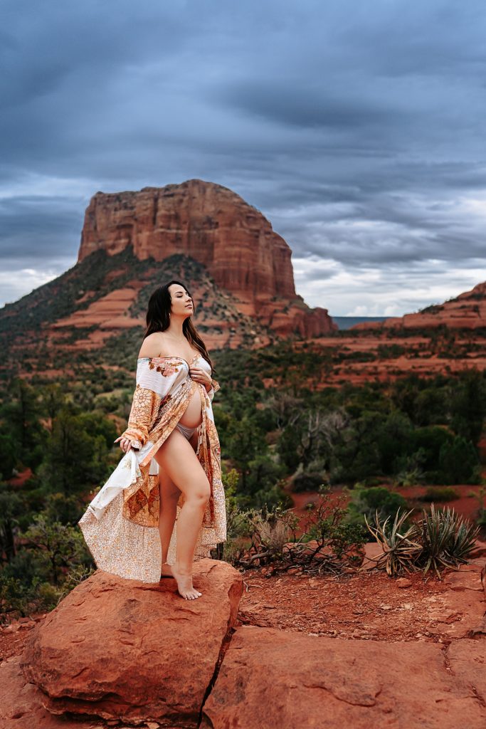Pregnant woman in a flowy, open dress, standing on an overlook with a gorgeous Arizona background in vibrant colors behind her.