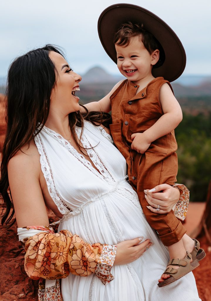 pregnant mother, holding her toddler who is wearing a hat, they are both laughing.