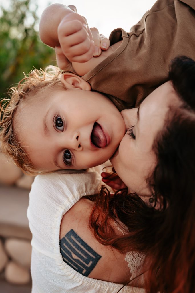 Toddler looking at the camera while mom holds him up to kiss his cheek.