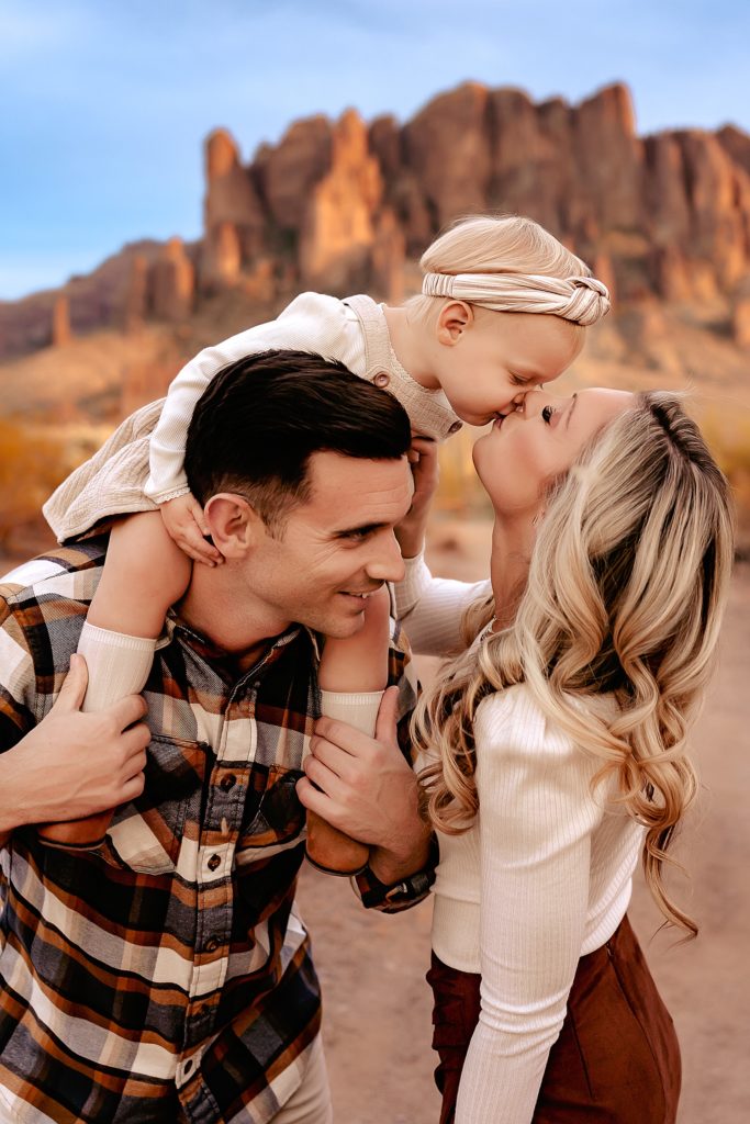 Mom kissing her toddler who is sitting on the father's shoulders for their family photo session in Phoenix. 
