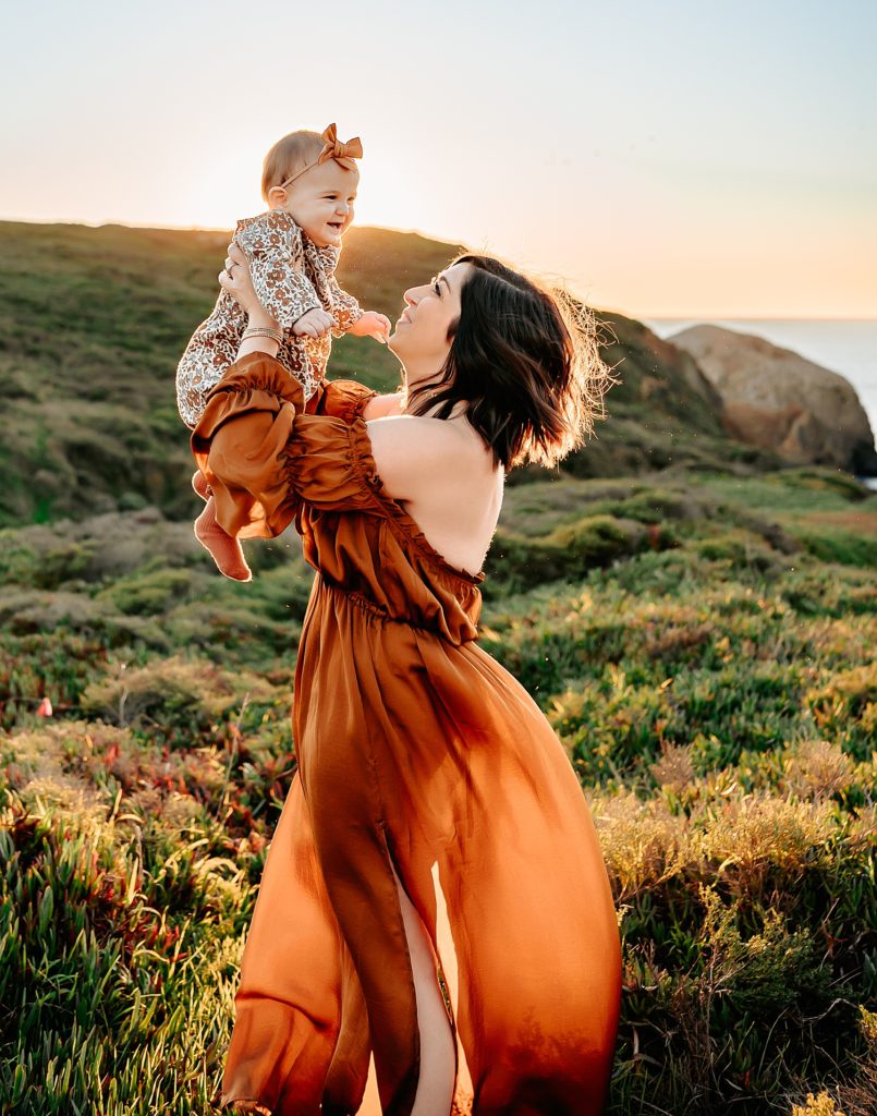 Mom in rust dress holding up her baby near the ocean with a green field behind them. 
