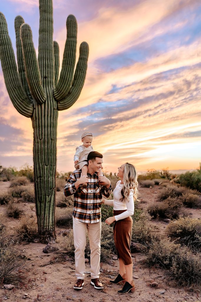 Baby toddler girl on dad's shoulders while mom laughs with her. They are standing next to a cactus with a gorgeous sunset above them. 