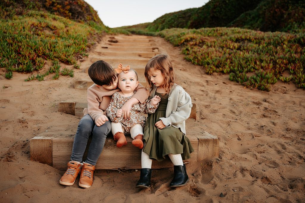 Three small children sitting on steps at the bottom of the stairs in the sand. 
