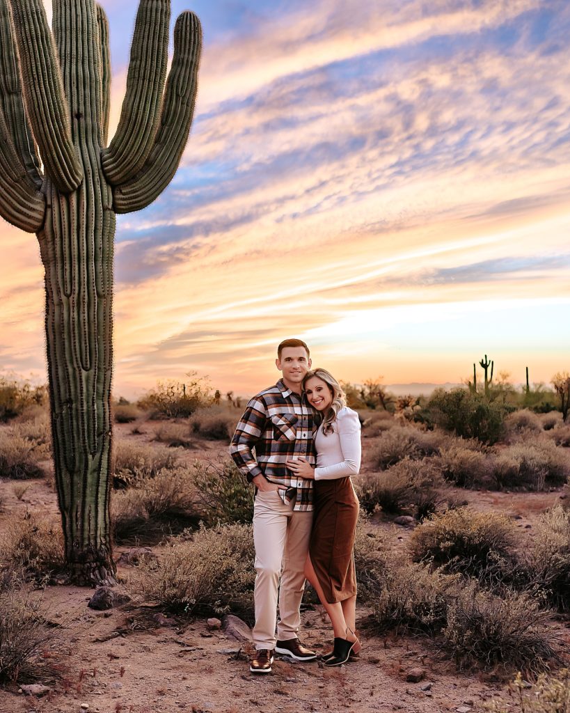 Blonde woman and man wearing a plaid shirt pose under a large cactus in Arizona with a beautiful sunset overhead. 