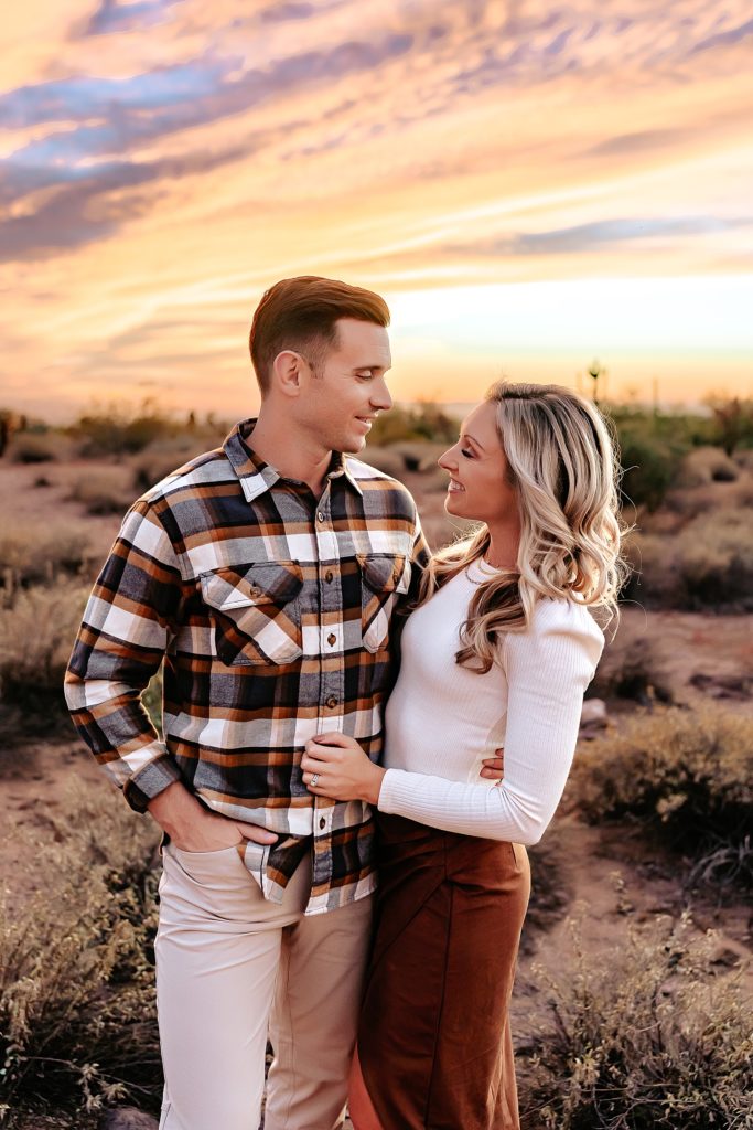 Woman with blonde hair and man wearing plaid embracing with a beautiful sunset overhead.