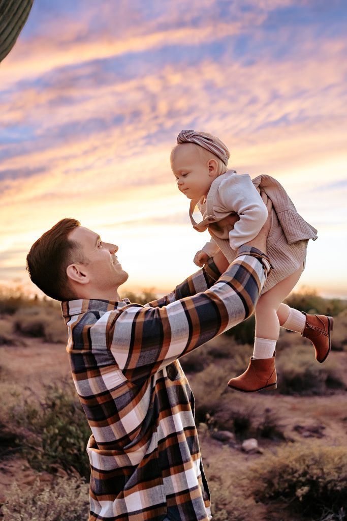 Dad throwing his baby toddler up in the air with a purple and orange sunset behind them. 